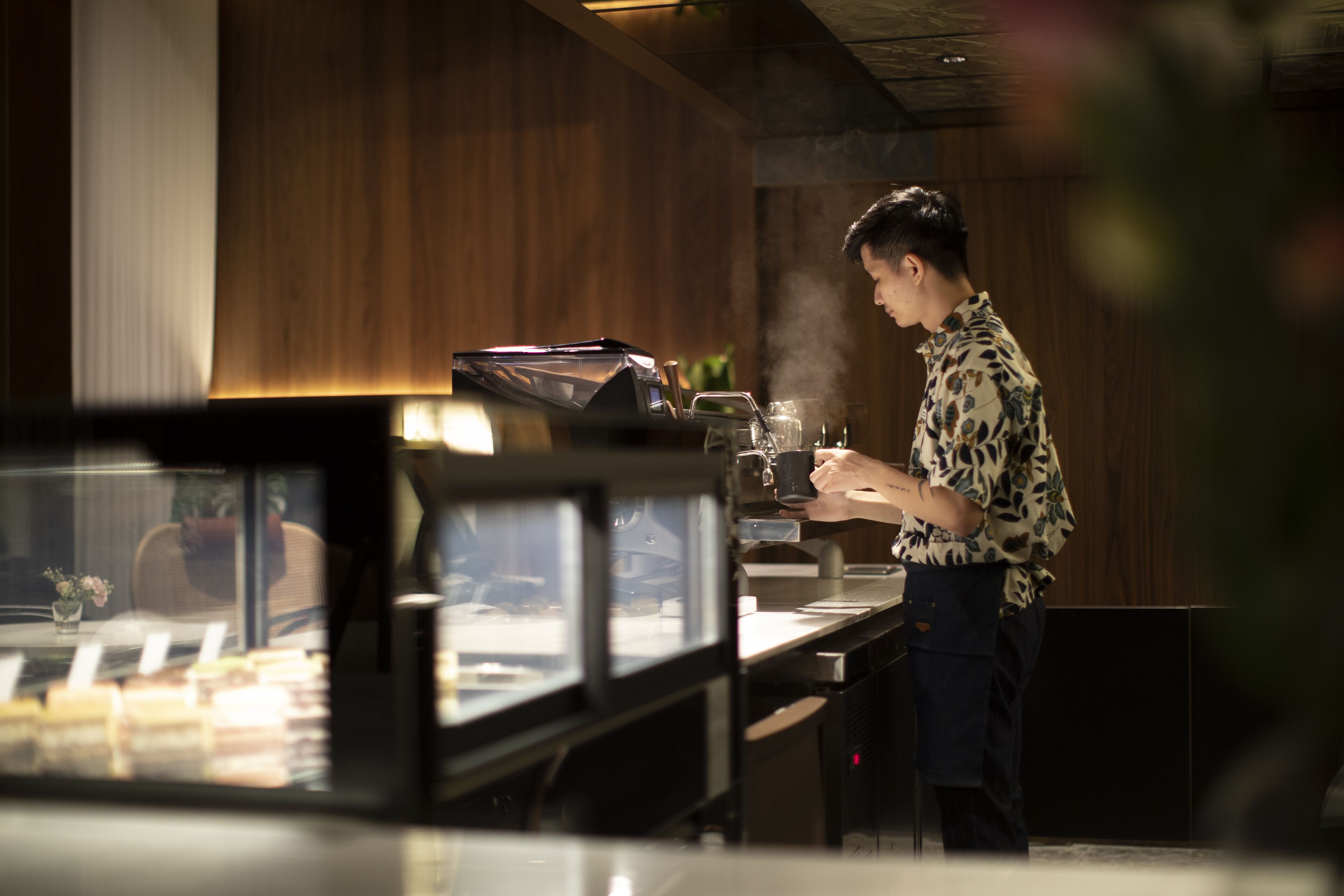 A person in a patterned shirt preparing a drink at a bar with a steam vent, a display case with items, and wooden paneling in the background.