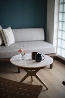 Una mesa redonda con un jarrón de flores y una taza, un sofá al fondo y una ventana con persianas.