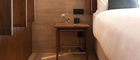 A hotel room with a wooden floor, a bed with white and teal pillows, a nightstand with a vase of flowers and a black mug, and a pair of shoes on the floor.