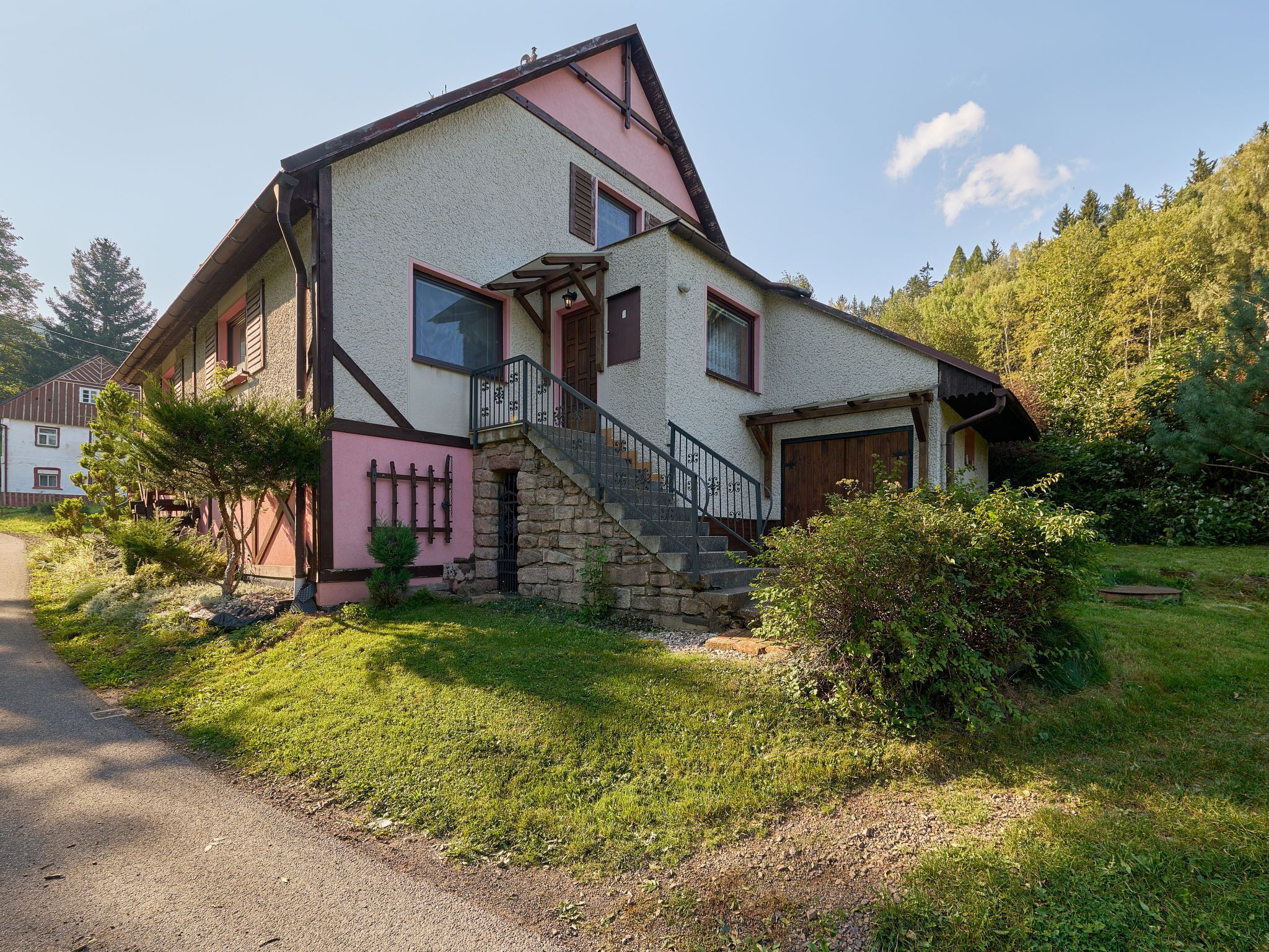 Une maison à deux étages avec un escalier en pierre menant à la porte d’entrée, le tout entouré de verdure.
