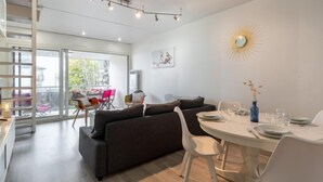 Una sala de estar moderna con un sofá gris oscuro, sillas blancas y una mesa de comedor preparada para cuatro personas. Una escalera conduce a un nivel superior, y un gran ventanal ofrece vistas de la vegetación exterior.
