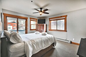 Une chambre à coucher comprenant un lit, un ventilateur de plafond, un téléviseur et des fenêtres avec des cadres en bois.