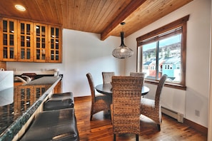 Un interior de madera con comedor, que incluye sillas de mimbre y una mesa, una cocina con armarios de madera, y un ventanal con vistas a edificios.