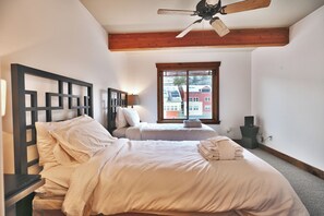 A hotel room with two beds, a ceiling fan, a window with a view of buildings, and a bedside table with a lamp.