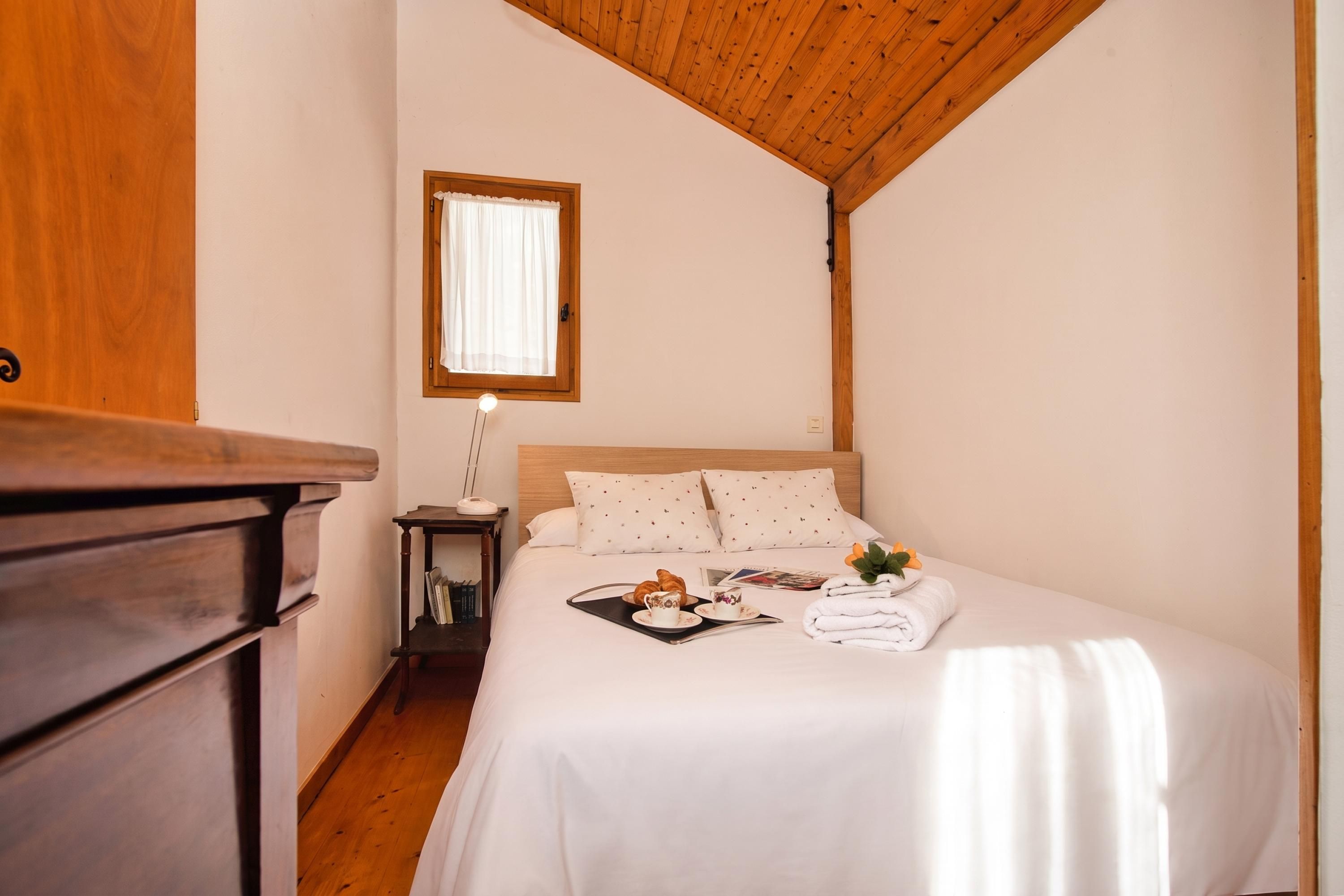 Ein Hotelzimmer mit Holzboden, einem Bett mit weißer Bettwäsche, einem Nachttisch mit Lampe und einem Fenster mit Vorhängen.
