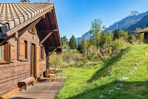 A wooden cabin with a sloped roof, a deck with outdoor seating, and a mountainous backdrop.