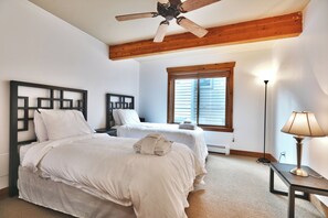 Une chambre avec deux lits, un ventilateur de plafond, une fenêtre avec des stores, une lampe sur pied et un radiateur.