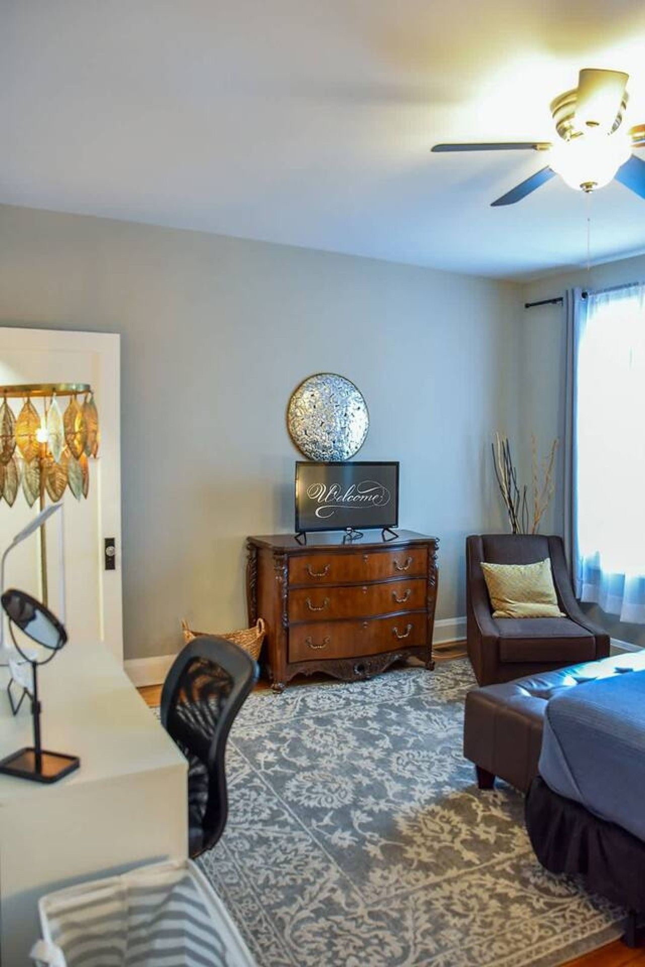 A hotel room with a bed, a desk, a dresser, a chair, a ceiling fan, and a decorative wall piece.