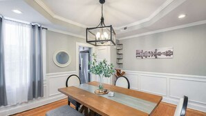 Un comedor con mesa de madera, bancos, lámpara colgante, planta en maceta y espejo montado en la pared.