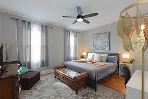 Un dormitorio con cama, mesitas de noche, escritorio, silla, ventilador de techo y ventana con cortinas.