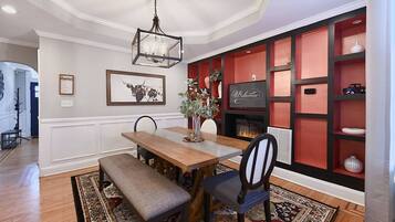 A dining area with a wooden table, bench, chairs, a chandelier, a fireplace, and a red shelving unit with decorative items.