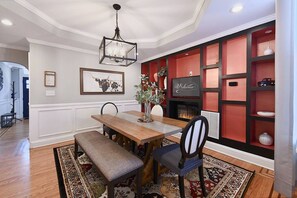 Un comedor con mesa de madera, banco, sillas, una araña, chimenea y una estantería roja con objetos decorativos.