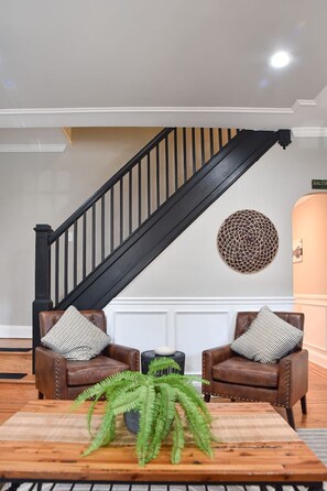 Una escalera con barandillas negras, dos sillones de cuero con cojines estampados, un adorno circular en la pared y una mesa de centro de madera con una planta verde.