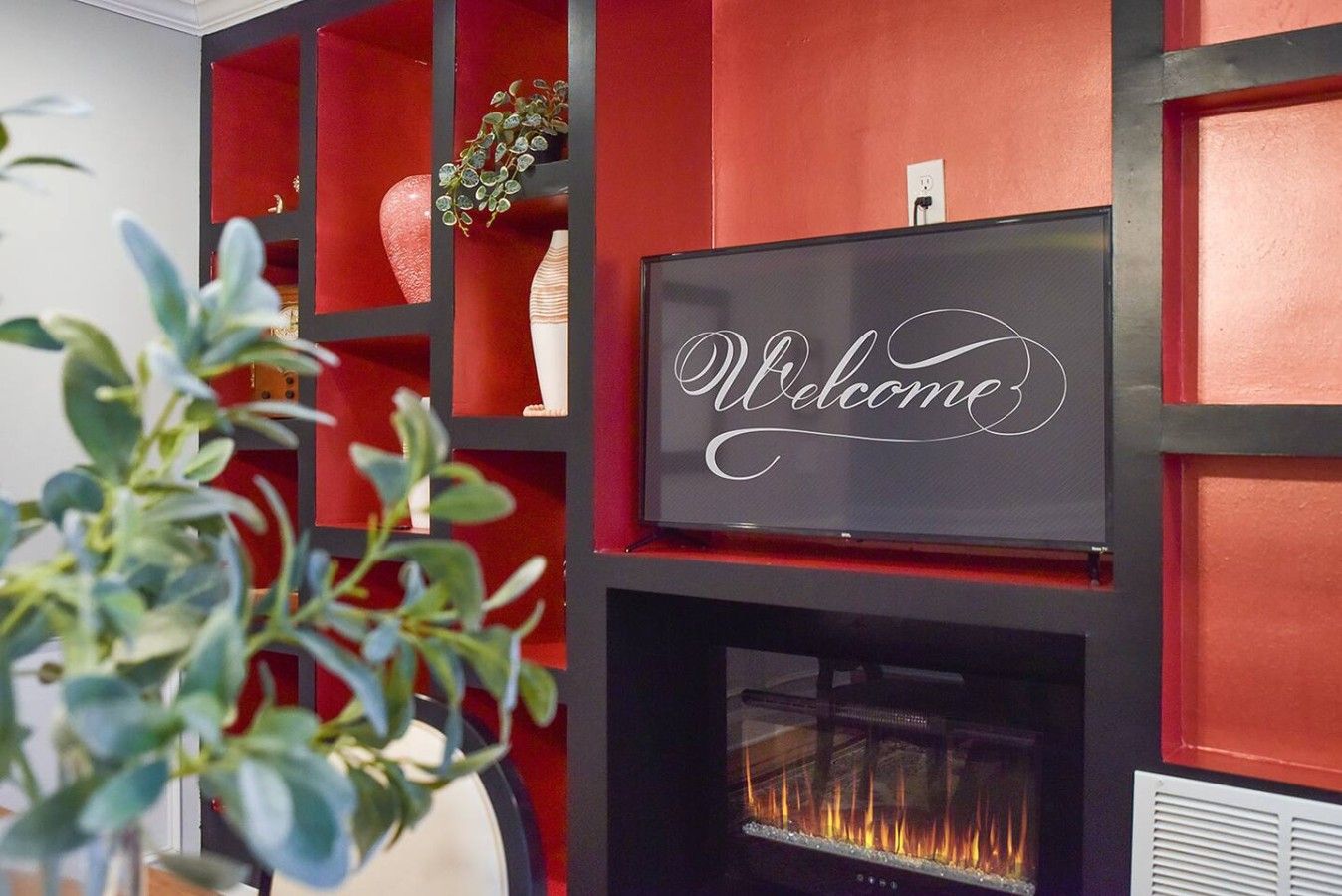 A fireplace with a 'Welcome' sign above it, surrounded by red shelving with decorative items.