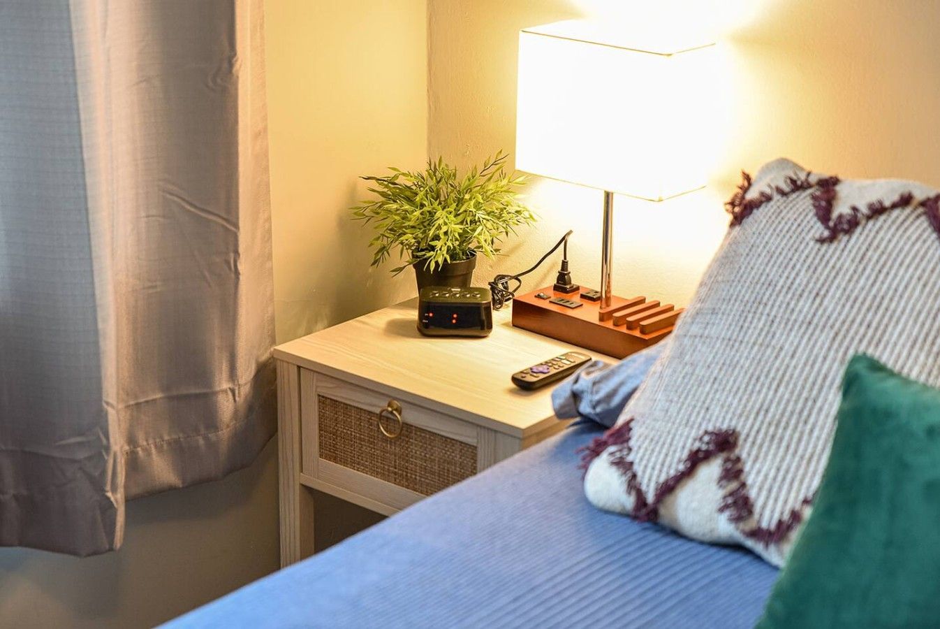 Un lit bien fait, avec une couverture bleue et un oreiller, une table de chevet avec une lampe, une petite plante et une télécommande.