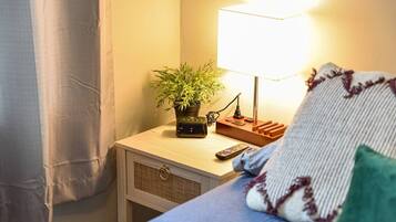 Un lit bien fait, avec une couverture bleue et un oreiller, une table de chevet avec une lampe, une petite plante et une télécommande.