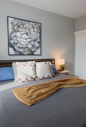 A bedroom with a bed, pillows, a blanket, a nightstand, a lamp, and a wall-mounted painting.