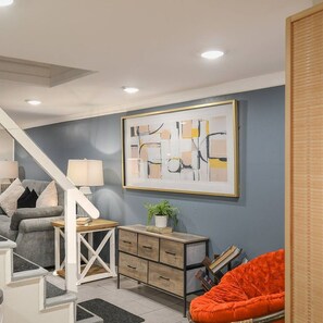 A modern living room with a grey sofa, a wooden side table, a bright orange chair, and a framed abstract painting on the wall.