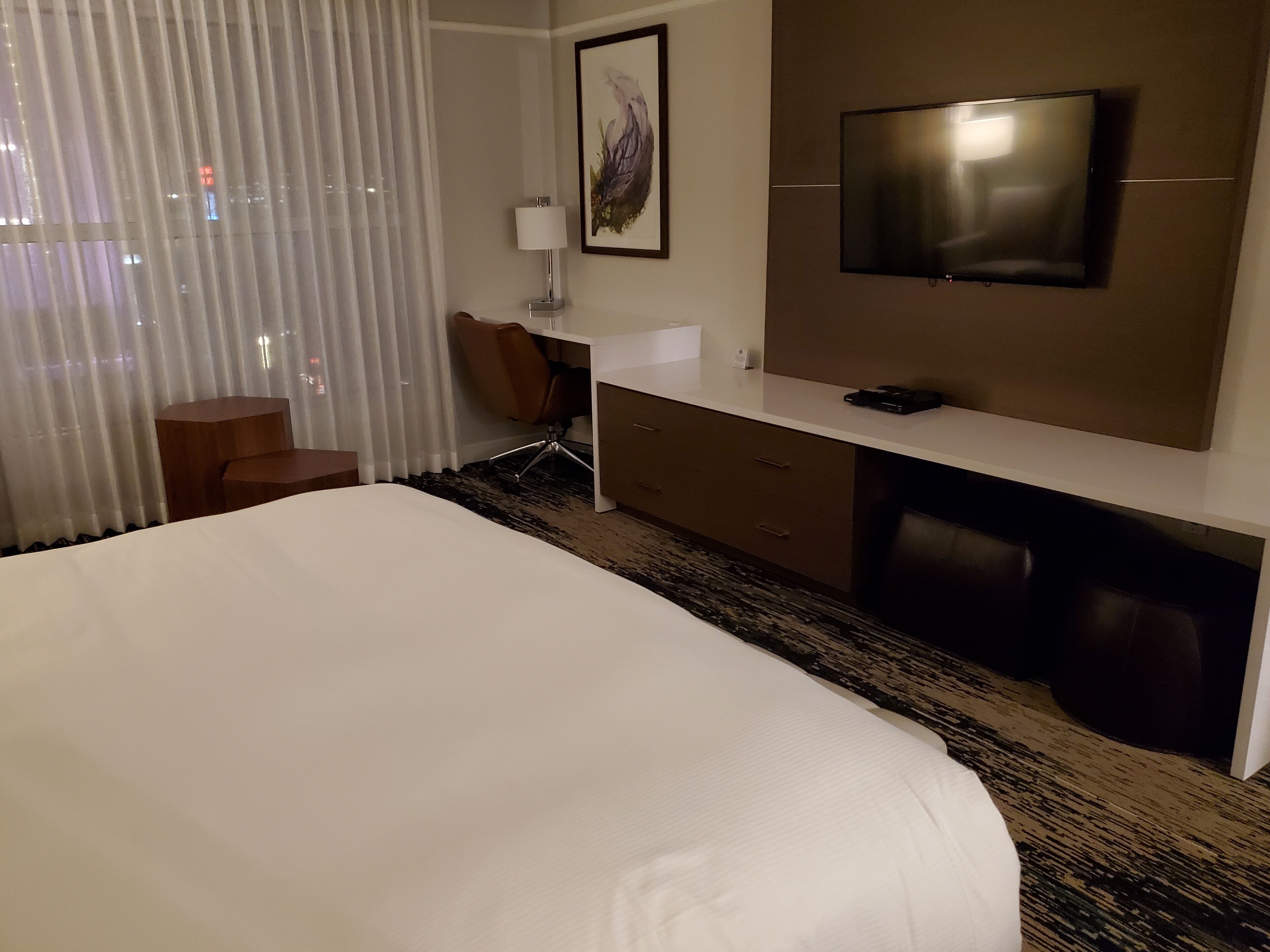 Ein Hotelzimmer mit einem großen Bett, einem Schreibtisch mit Stuhl, einem Fernseher und einem Fenster mit Vorhängen.