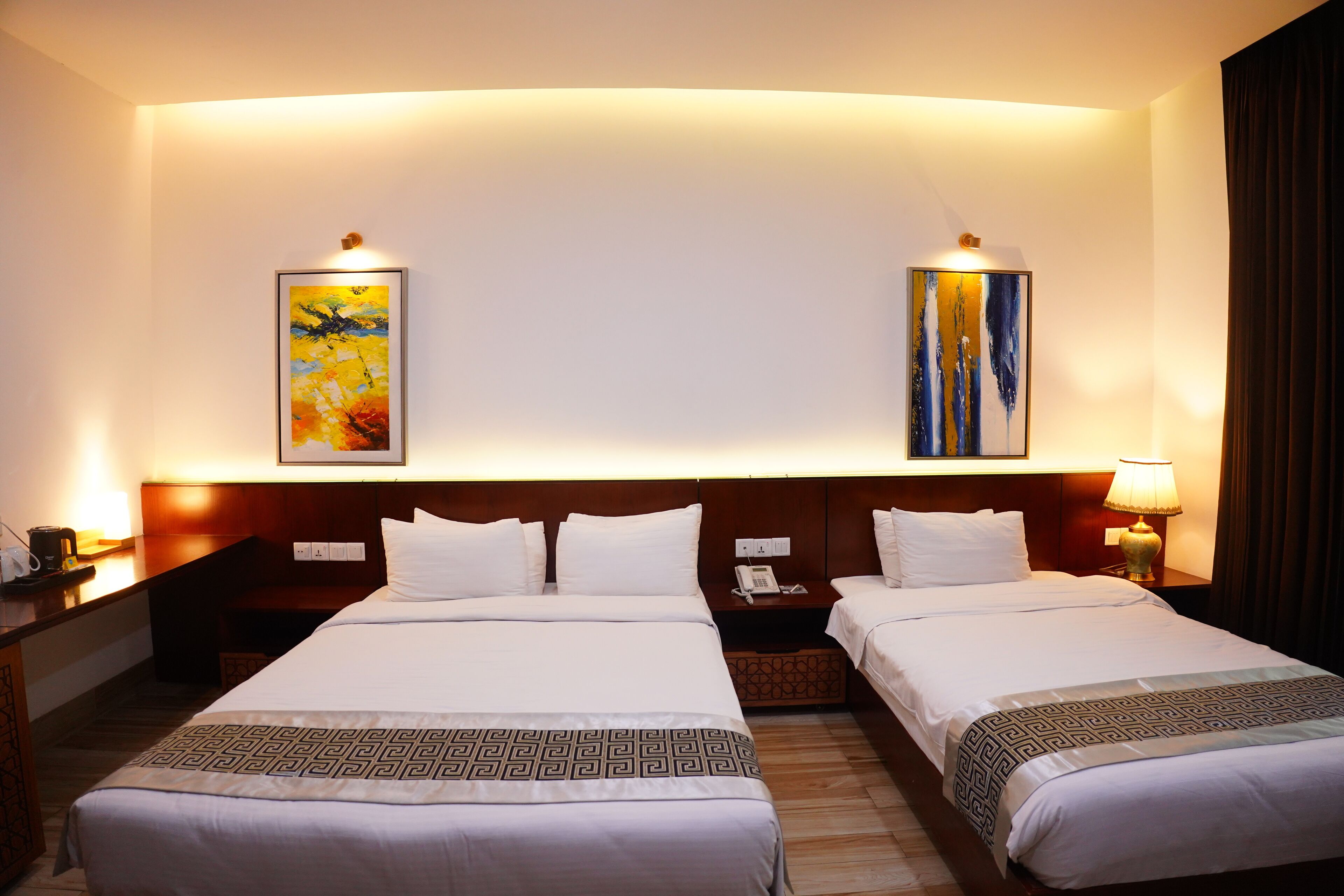 A hotel room with two beds, a wooden headboard, a nightstand with a lamp, and artwork on the walls.