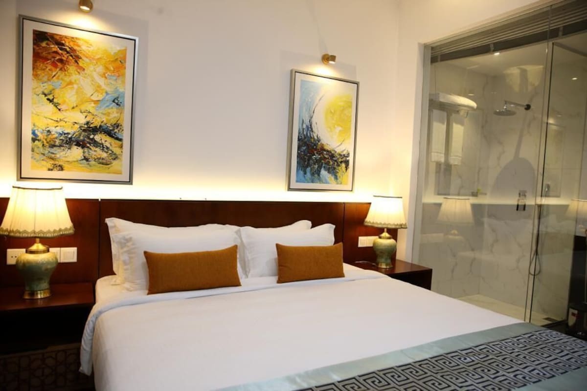 A hotel room with a large bed, two wall-mounted paintings, a glass shower enclosure, and two bedside lamps.