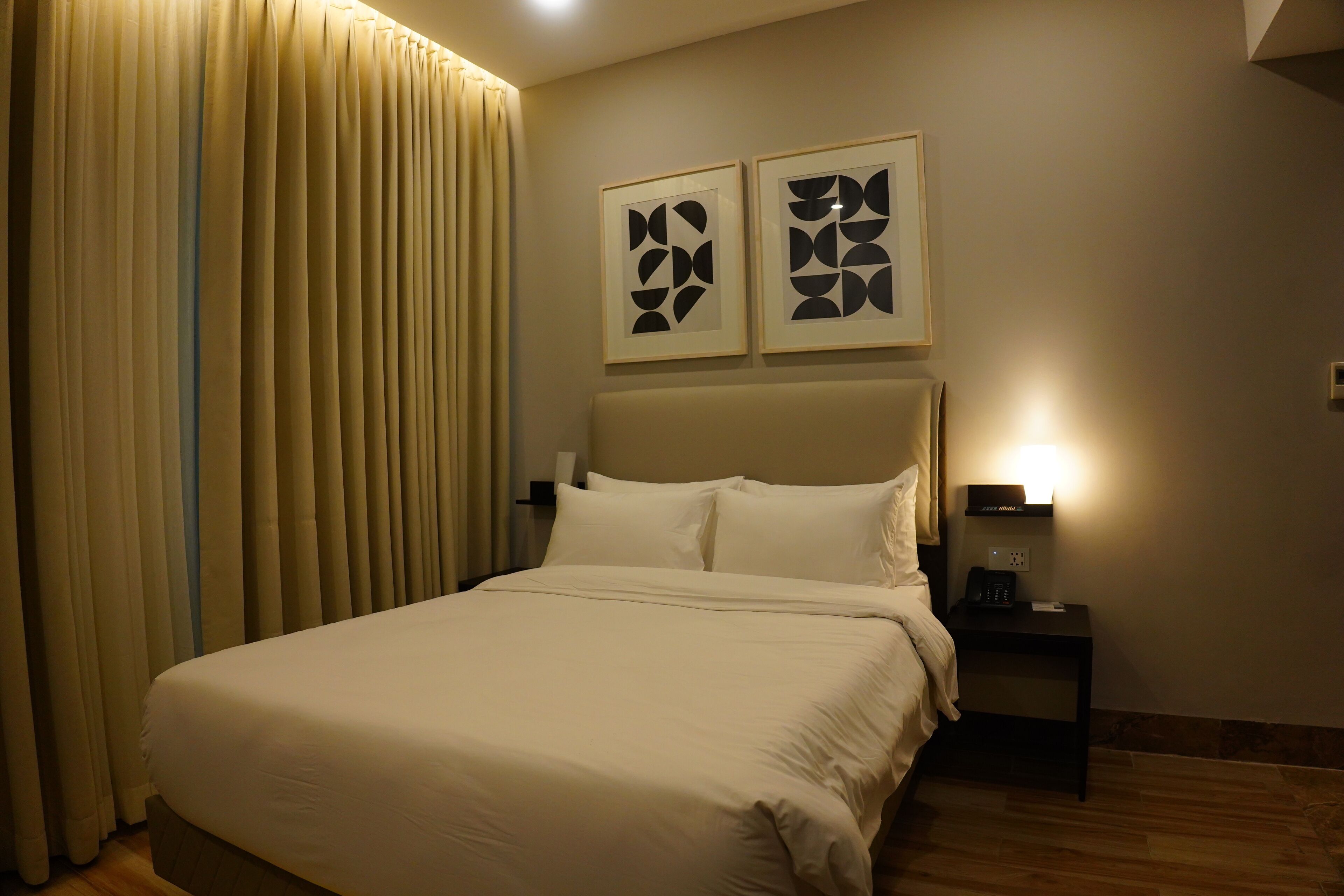 Una cama bien hecha con cabecera, una mesita de noche con lámpara y dos cuadros abstractos enmarcados en la pared.