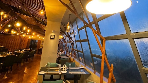 A restaurant with a modern interior, featuring a long table with chairs, a large window with a view, and hanging lights. - Empyrean Hotel (Dhaka)