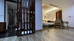 A hotel room with a modern design, featuring a wooden floor, a bed with a white headboard, a brown armchair, a small table, and a decorative ceiling light. - Empyrean Hotel (Dhaka)