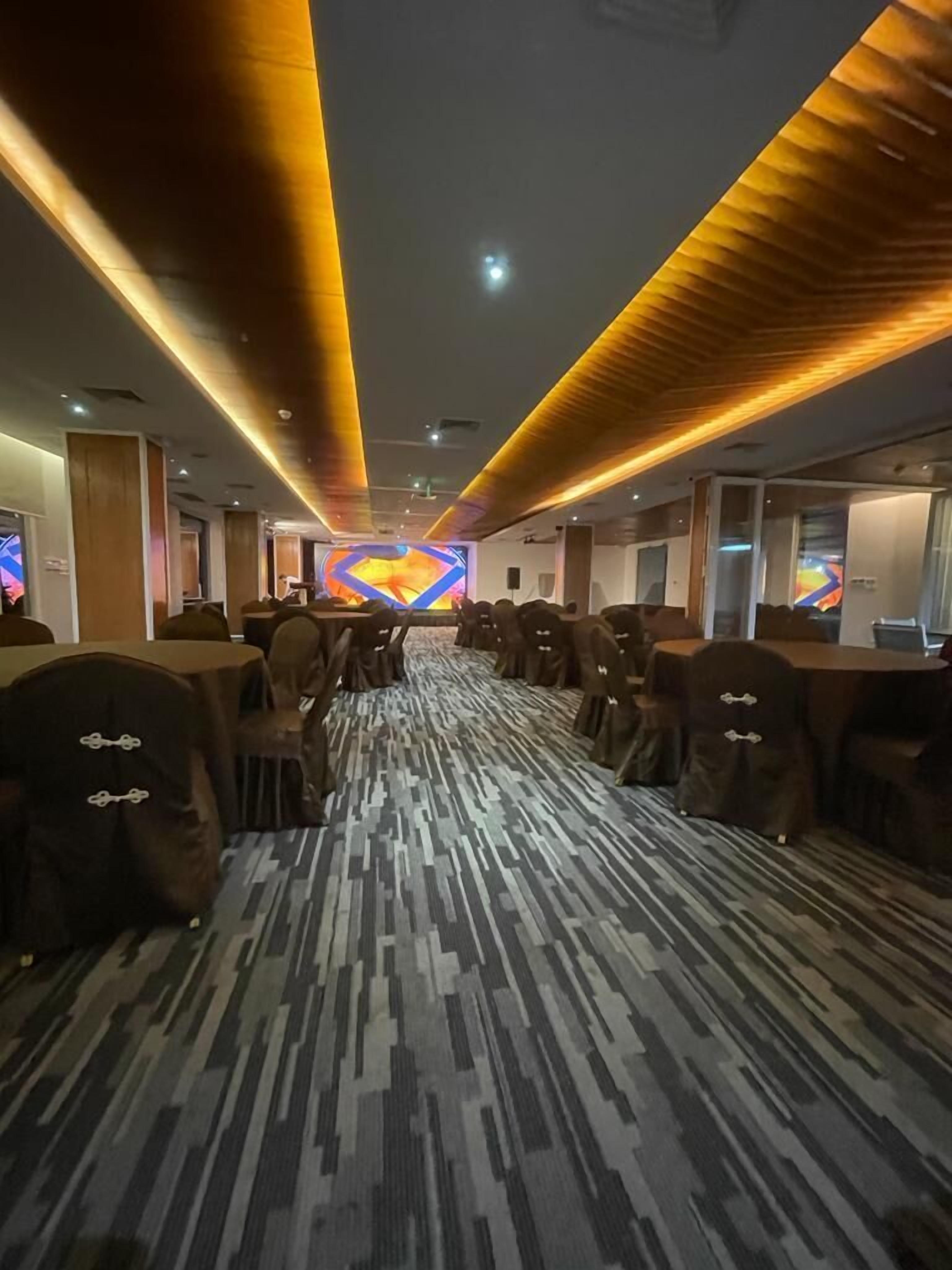 Corredor de hotel com fileiras de mesas e cadeiras, carpete estampado e uma tela digital grande no final.
