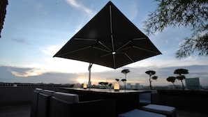 A rooftop terrace with a large umbrella, wicker furniture, and a view of the sunset over the city. - Empyrean Hotel (Dhaka)