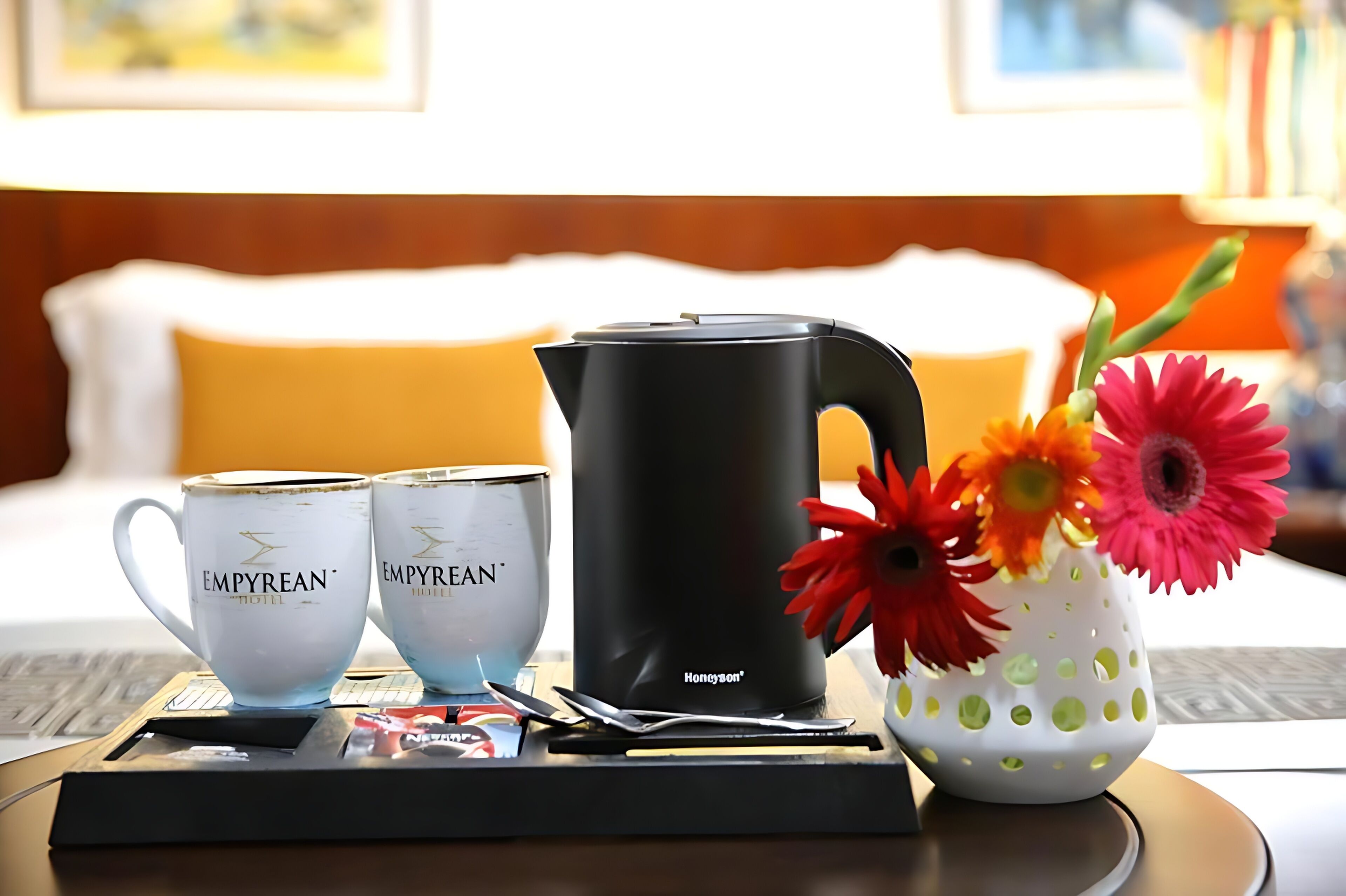 A hotel room with a bed, a tray table with two cups, a kettle, and a vase with flowers.