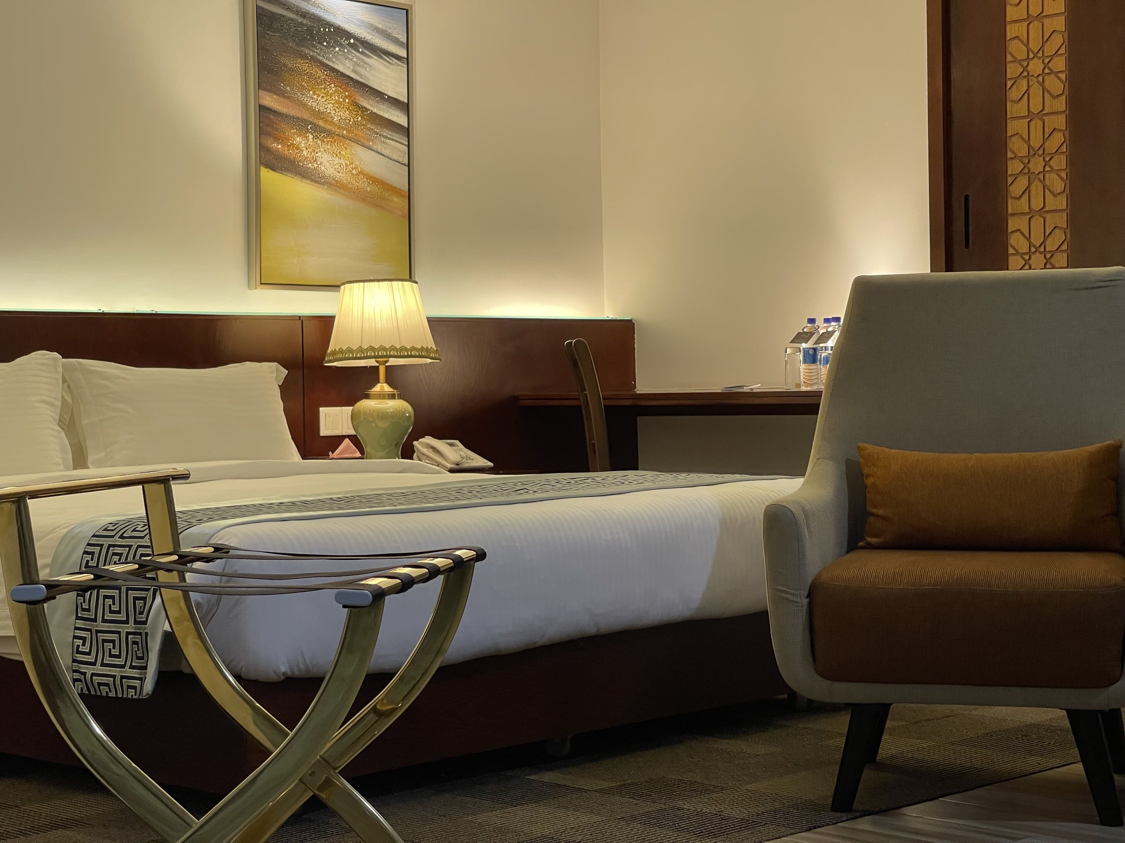 A hotel room with a bed, a chair, a desk with a lamp, and a wall-mounted artwork.