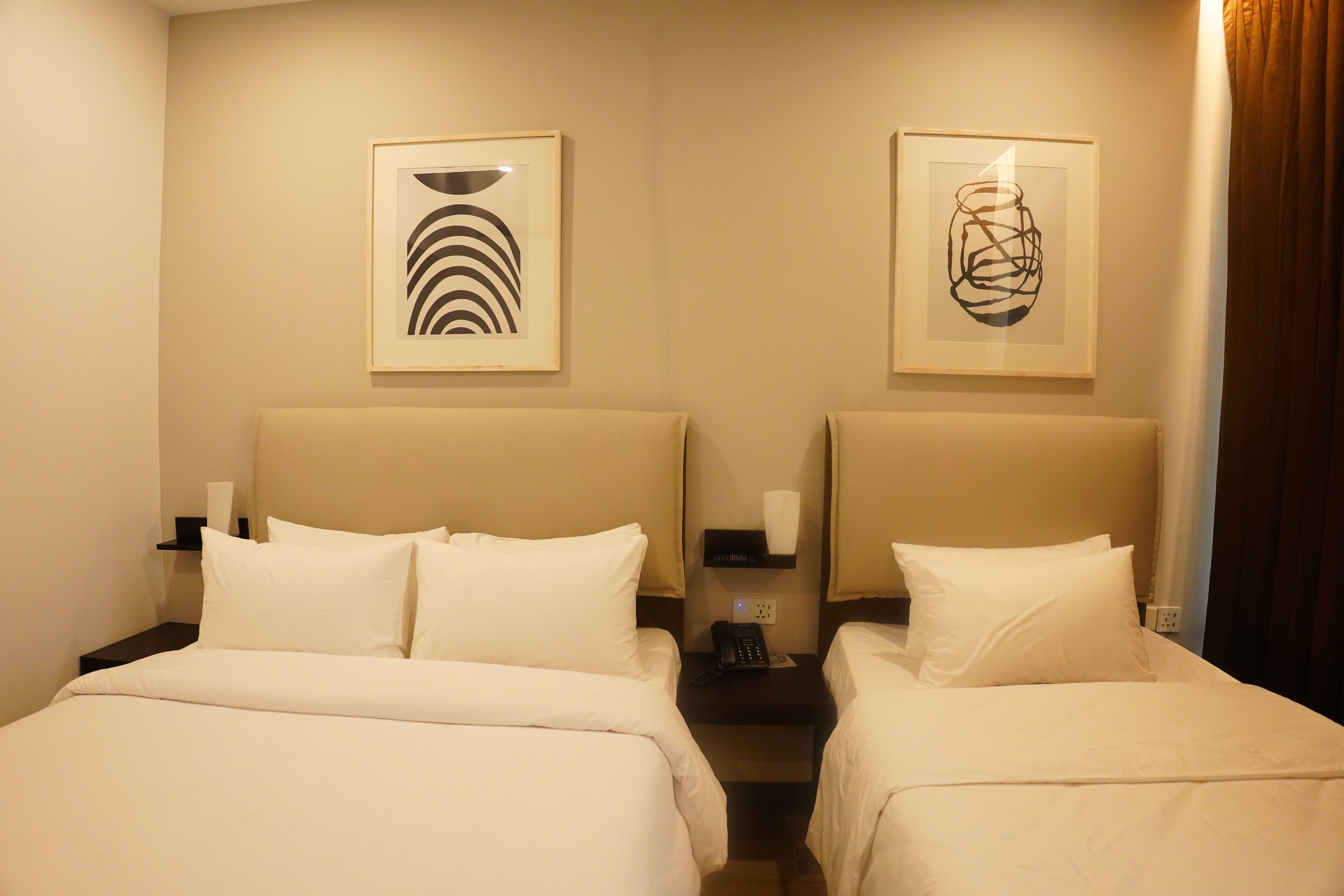 A hotel room with two beds, each with white bedding and headboards. There are framed abstract artworks on the walls above the beds.