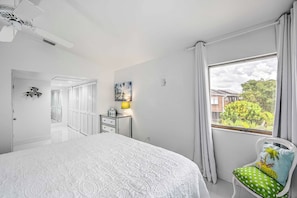 Un dormitorio con una cama grande, una ventana con cortinas, una silla con cojín, una mesita de noche con lámpara y una puerta que da a otra habitación.
