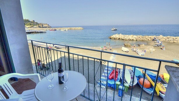 Un balcón con una mesa, dos copas de vino y una botella. El balcón tiene vista a una playa con sombrillas y sillas.