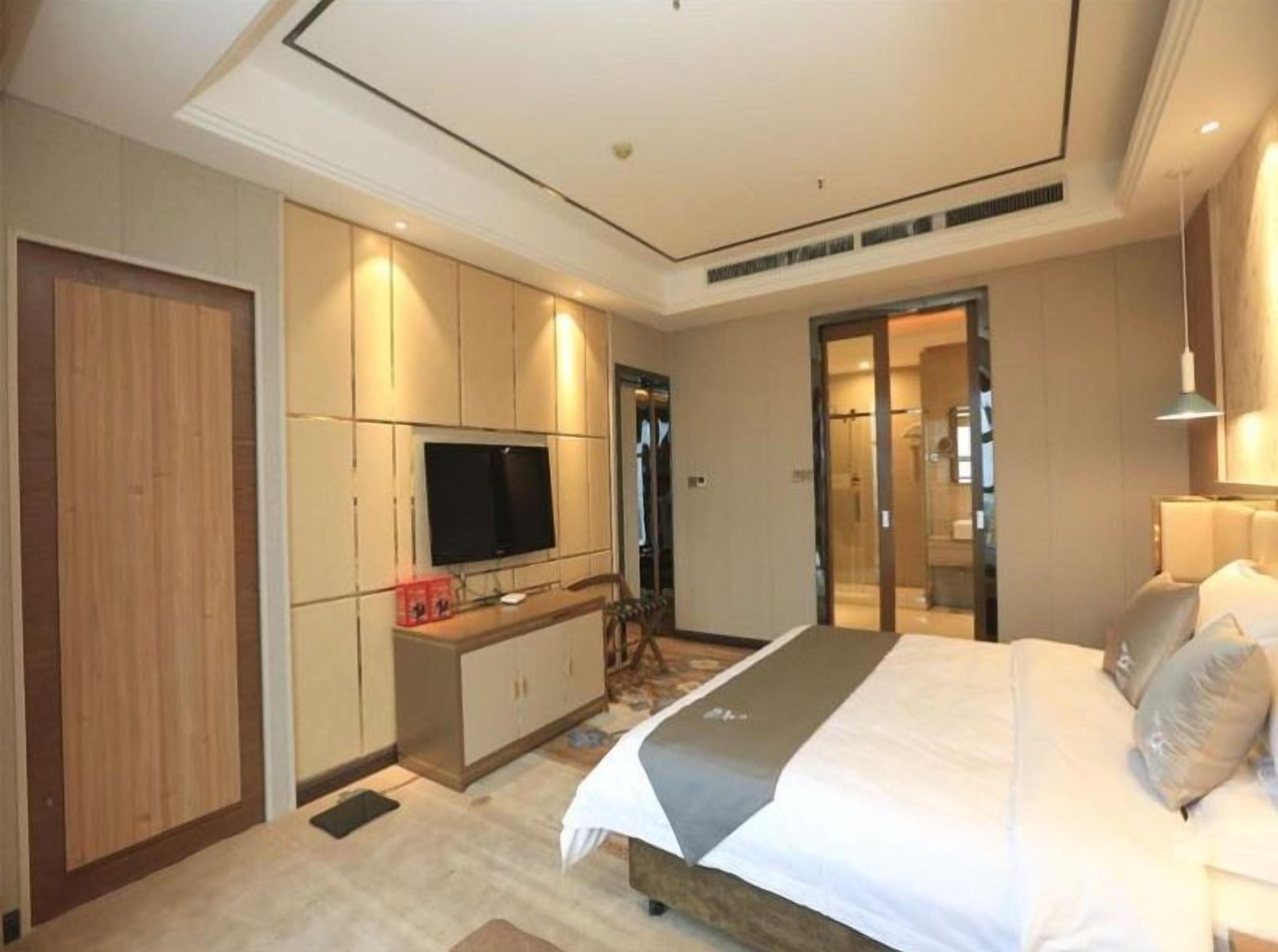 Una habitación de hotel moderna con una cama grande, un televisor montado en la pared, un vestidor y un baño con puerta de cristal.