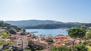 Un pueblo costero con edificios a lo largo del paseo marítimo, un puerto deportivo con barcos y colinas al fondo.