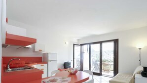 Una cocina compacta con encimera roja, un lavamanos y un refrigerador. Hay una zona de comedor con una mesa y sillas, y un amplio ventanal corredero que da a un área exterior con vistas a árboles y un edificio.
