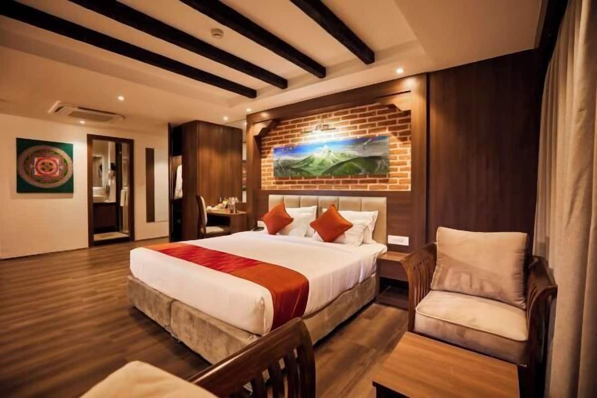 A hotel room with a large bed, a wooden headboard, a painting above the bed, a wooden floor, a chair, and a nightstand.