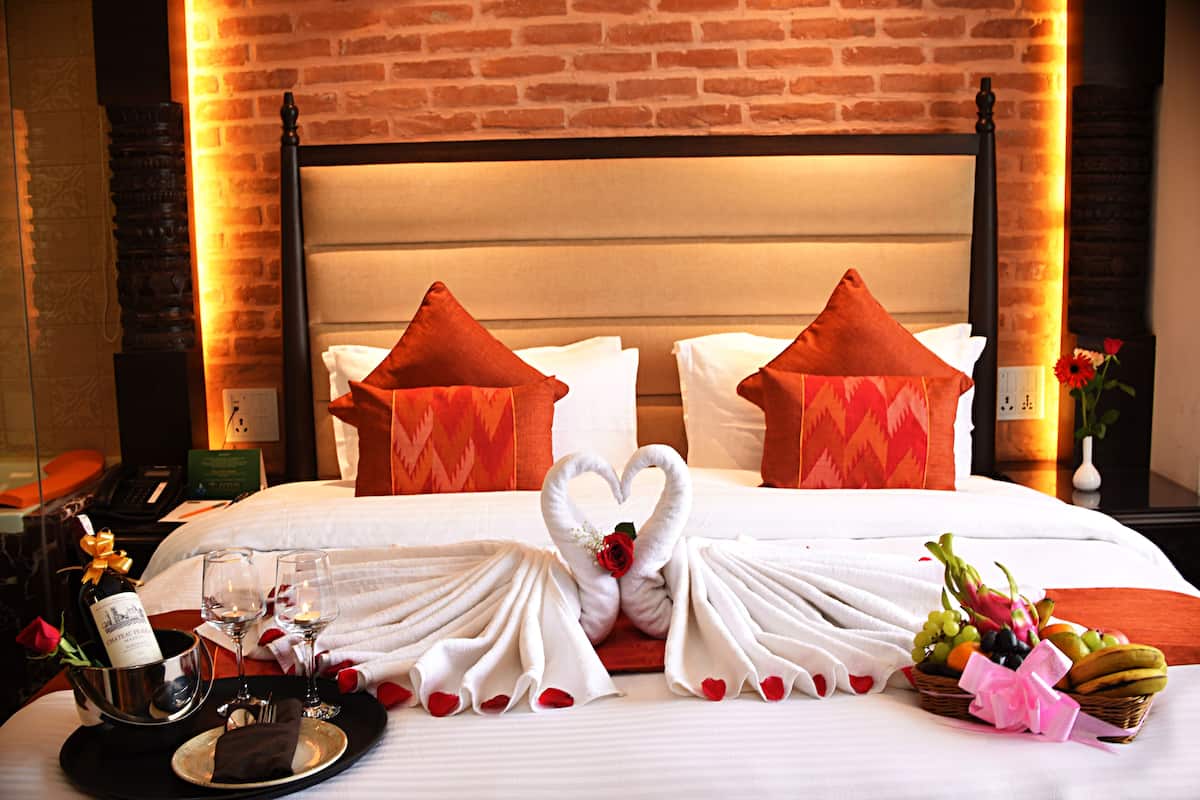A hotel room with a brick wall, a bed with white bedding, red and white pillows, a swan-shaped towel arrangement, a tray with a bottle of champagne, glasses, and a fruit basket.