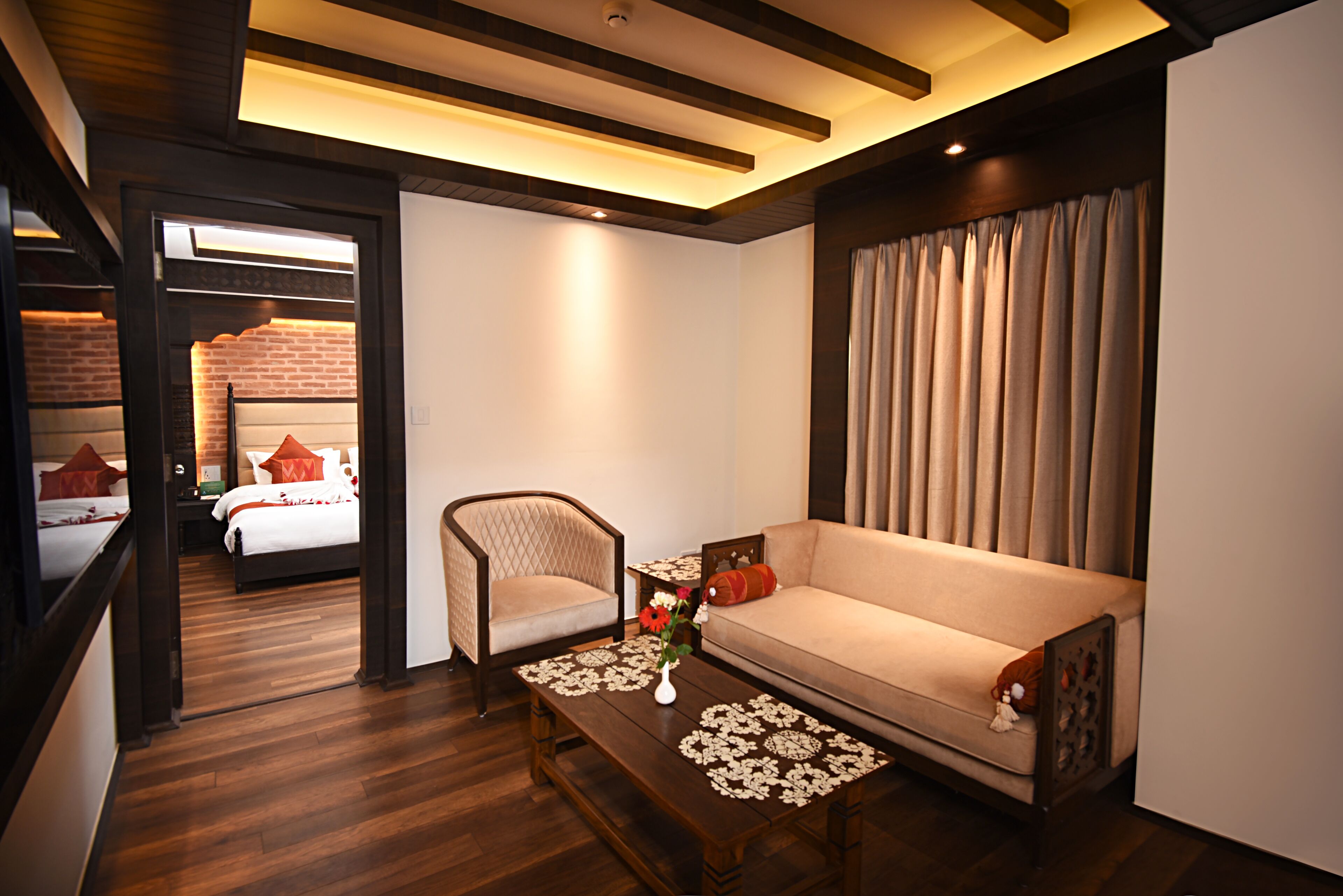 A hotel room with a sofa, a chair, a coffee table with a vase, a bed with red pillows, and a brick wall.