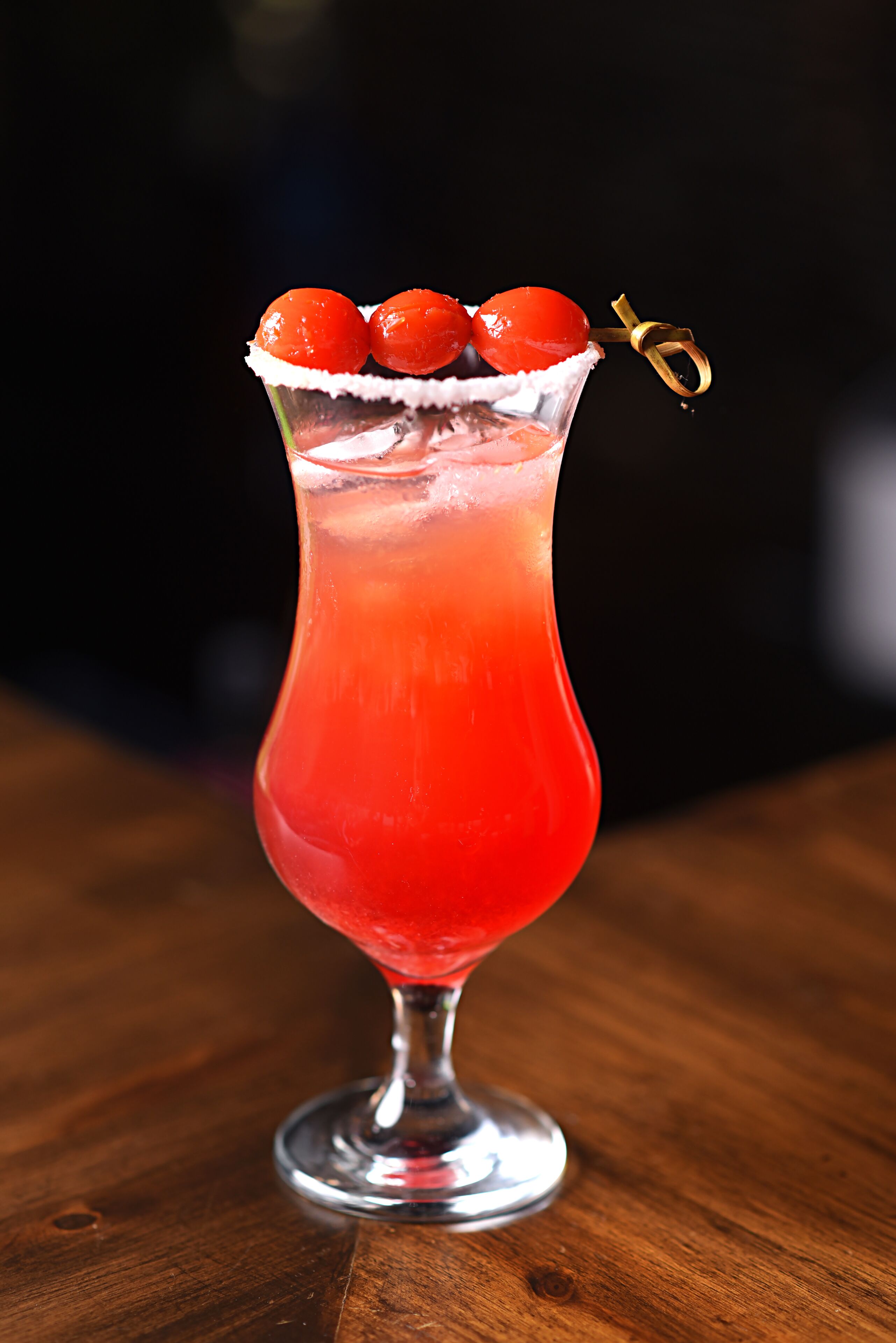 Un cóctel en un vaso alto con un líquido rojo y cerezas en el borde.