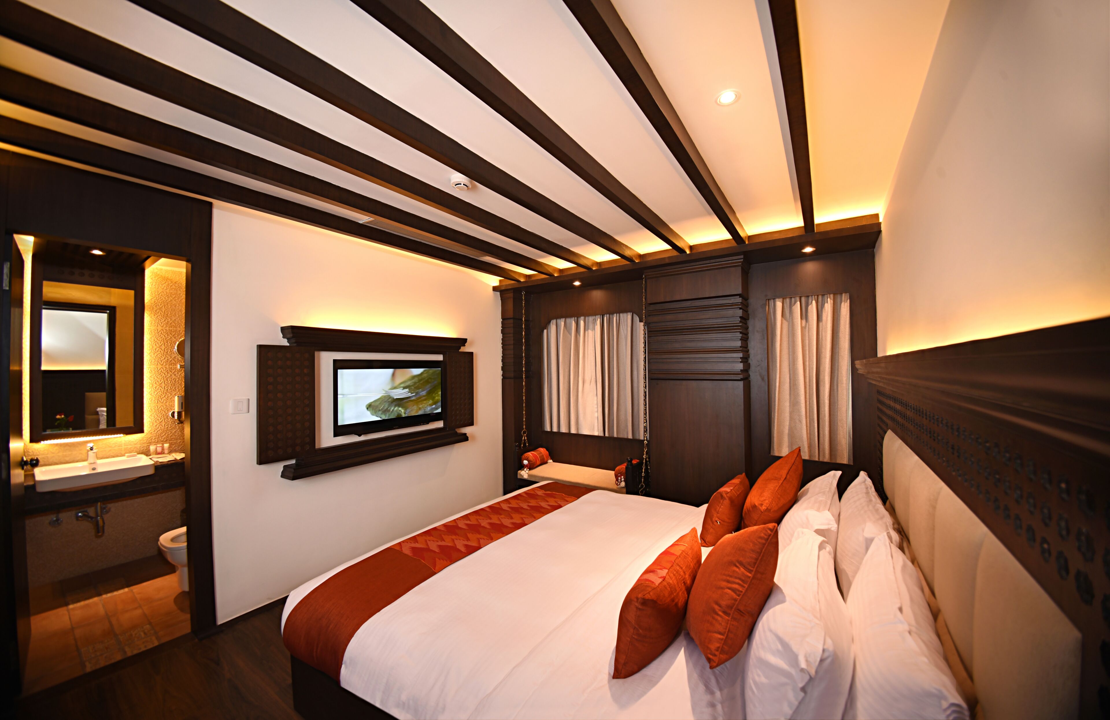 A hotel room with a large bed, a television mounted on the wall, a bathroom with a sink and toilet, and a wooden ceiling with exposed beams.