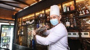 A bartender in a white uniform and hat preparing a drink at a bar with shelves of liquor bottles. - Kantipur Village (Kathmandu)