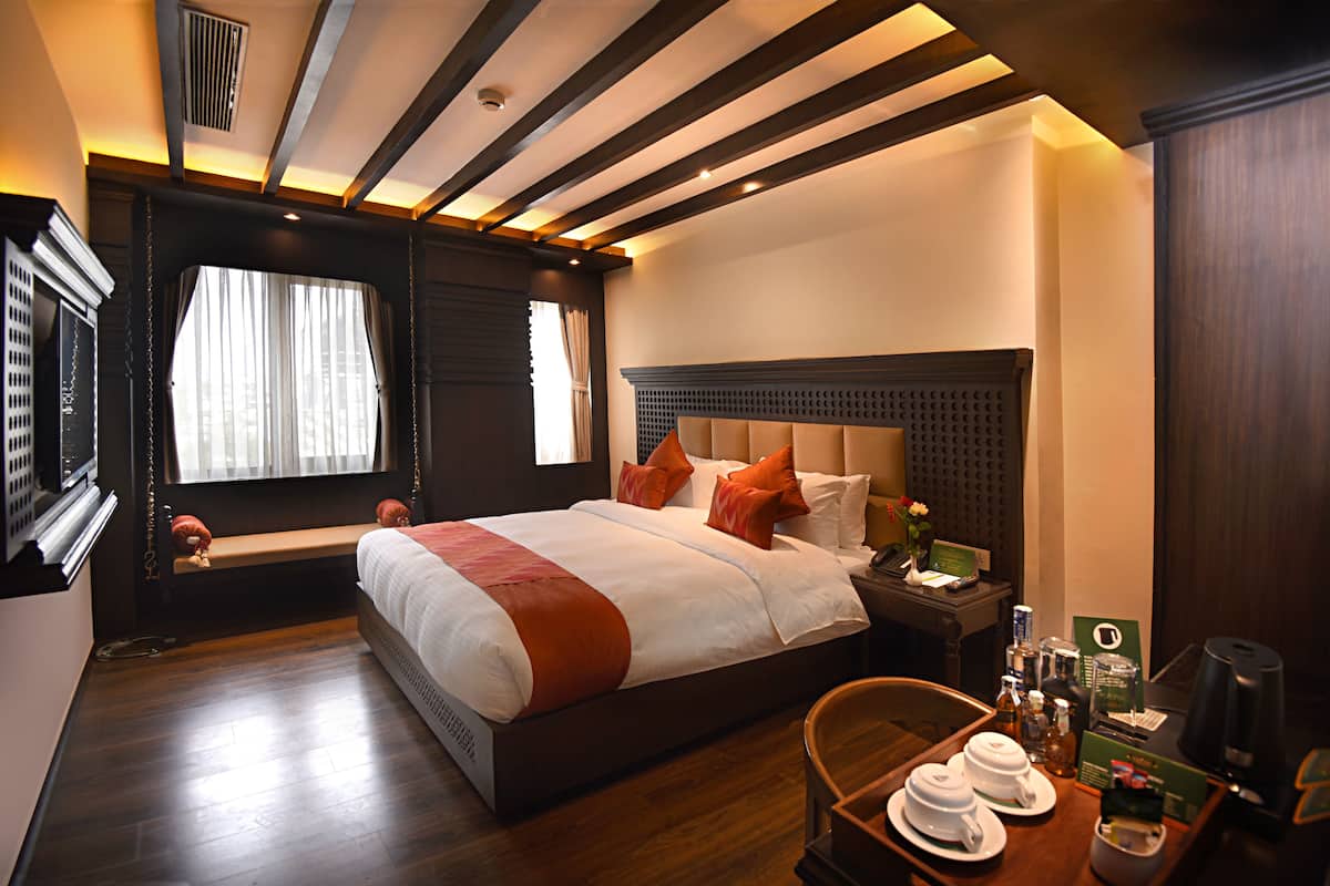 A hotel room with a large bed, a bedside table with a lamp, a chair, and a small table with tea cups and a tray.
