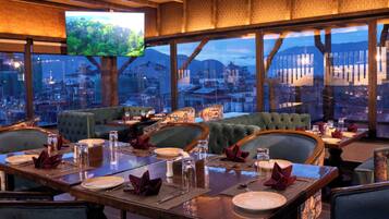 A restaurant with a high ceiling, large windows, and a city view. Tables are set with plates, glasses, and napkins.