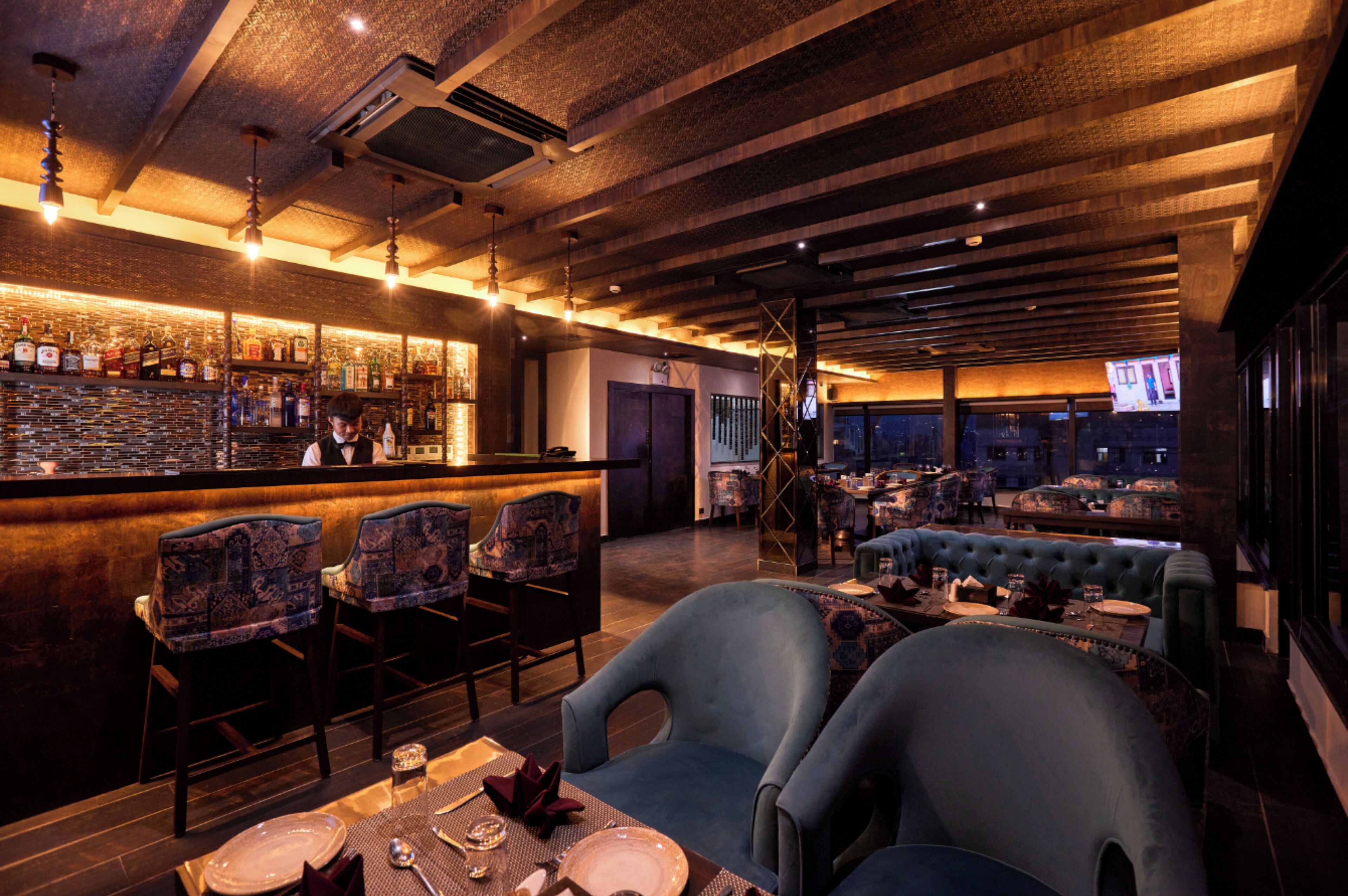 A bar area with a wooden ceiling, bar stools, and a counter with bottles. There are tables with chairs and a sofa in the seating area.