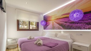 A bedroom with a large bed, a nightstand, a wall-mounted lamp, and a window with a view of trees.