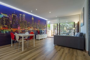 Een moderne woonkamer met een stadsgezicht muurschildering, een eetgedeelte met rode stoelen, een grijze bank en een witte tv-meubel.