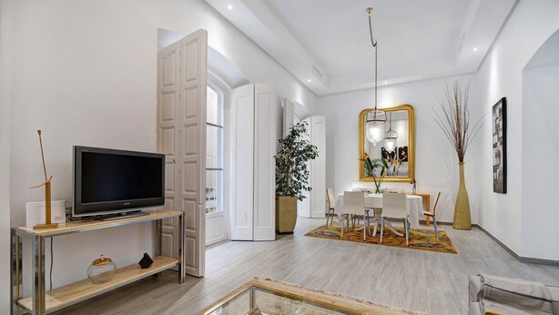 A modern living room with a flat-screen TV, a glass coffee table, a sofa, and a dining area with a white table and chairs.